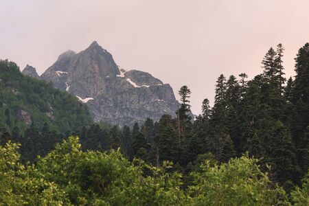 View of a high steep rocky mountain partially covered with snow against a dark cloudy sky with...