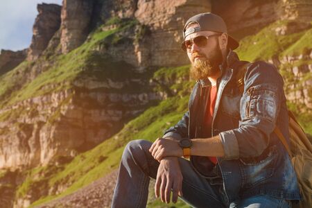 Close-up of a bearded man in jeans clothes in sunglasses and a cap with a backpack sitting at the...