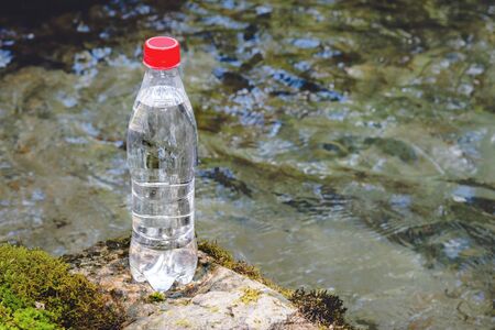 A plastic bottle with a red cap with fresh drinking water against a background of green forest and the waters of a mountain river stands on a stone with moss. The concept of pure natural mountain water.の写真素材