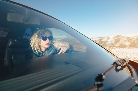 Attractive Caucasian girl in sunglasses and a leather jacket sits at the wheel of a sports retro car...