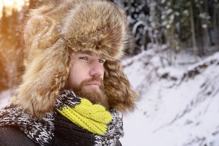 Portrait of an attractive manly man with a mustache and beard in a winter forest. Stylish tourist in a fur hat and a bright scarfの写真素材