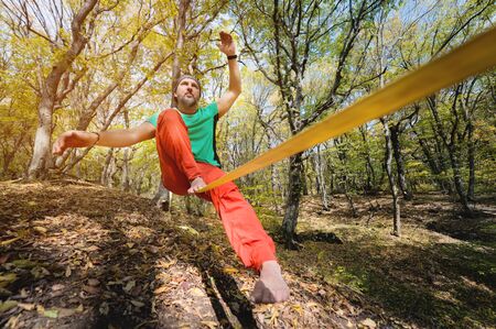 Wide angle male tightrope walker balancing while sitting barefoot on slackline in autumn forest. The concept of outdoor sports and active life of people agedの写真素材