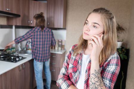 A young girl is talking on a cell phone in the kitchen amid her husband or boyfriend washing dishes. Secrets in the family and treason hiding from each otherの写真素材