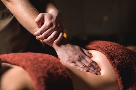 Close-up male hand of a professional physiotherapist masseur doing lumbar massage to a female patient in a massage luxury room with dim lightの写真素材