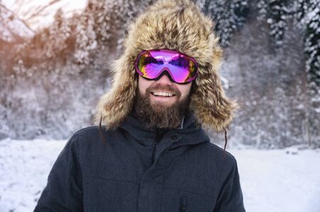 Close-up portrait of a bearded happy snowboarder skier in a ski mask with goggles and a fur big old-school hat on a background of a winter snowy mountainsの写真素材