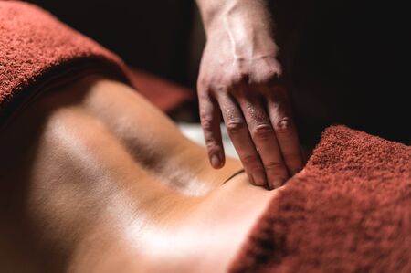Close-up male hand of a professional physiotherapist masseur doing lumbar massage to a female patient in a massage luxury room with dim light and a dark background with burning candles. The concept of luxury massage and health careの写真素材