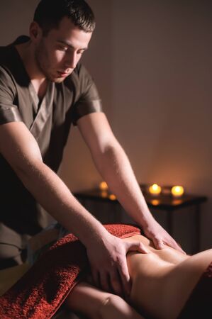 Professional premium massage in a dark atmospheric cabinet. Young man doing massage to a female client in a dark office on the background of burning candlesの写真素材