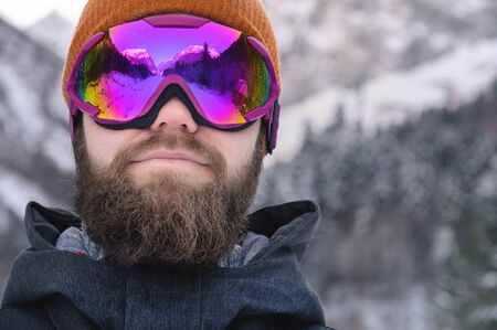 Close-up portrait of an attractive young bearded man in a knitted winter hat and ski mask with goggles on his face in the winter season. Portrait of a sportsman on a background of a winter forest with reflection of mountains in a maskの写真素材