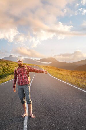 Stylish barefoot bearded male hitchhiker traveler in a hat and with a backpack walks along a suburban asphalt road in the mountains at sunset and stops the car with a hitchの写真素材