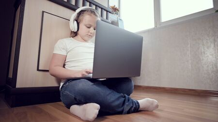 A smart little girl in white headphones with a laptop in her hands is pushing on the floor in her...