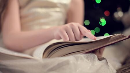 Close-up soft focus the hand of a little girl leads along the lines of a book while reading a New Years fairy tale amid the lights of a Christmas tree. The concept of a fabulous moodの写真素材