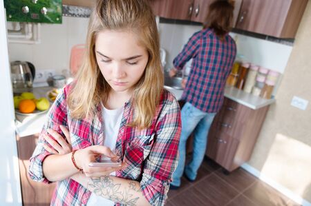 A girl is standing in the kitchen with a phone in her hands and is scamming a message surfing the Internet ordering food or writing a message to her loverの写真素材