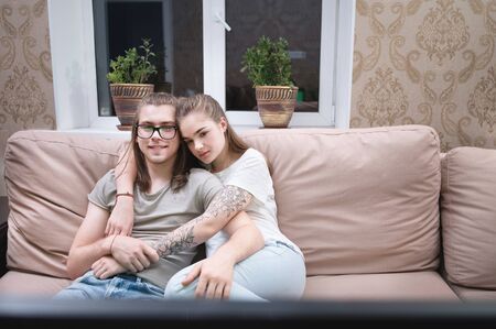 Attractive young long-haired couple hugging on a sofa at home in isolation watching tv. Concept of spending time with loved ones during a pandemicの写真素材