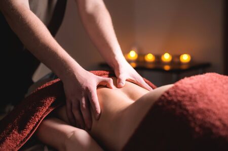A male professional massage therapist does back massage to a female client in a dark cozy medical room against a background of burning candles. Low key. The concept of premium massage and massage services.の写真素材