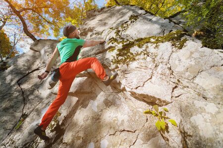 An aged bearded man is training in free climbing on a rock stone in the forest on a sunny day. The...