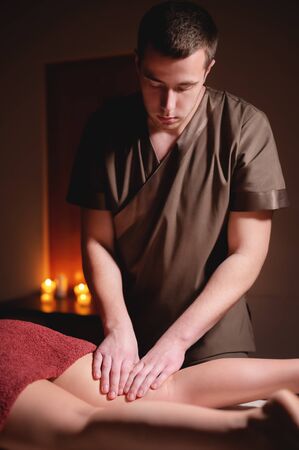 Close-up professional calf muscle massage to a female client by a male physiotherapist in a massage parlor in a dark office against the background of burning candlesの写真素材