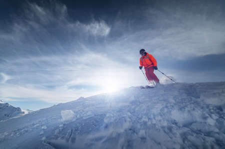An athlete male skier jumps from a snow-covered slope against the backdrop of a mountain landscape of snow-covered mountains on a sunny day. The concept of winter sports.の写真素材