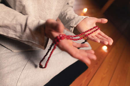 close-up man in clothes for practice and meditation sits in a lotus pose and holds red rosary to concentrate attention in a wooden room with dim lightの写真素材