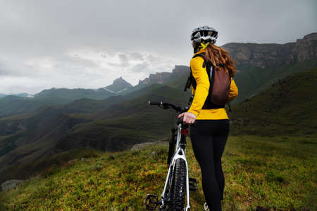 a girl in a helmet and with a backpack with a bicycle stands against the backdrop of epic mountains...