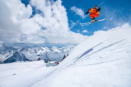 A male skier in an orange suit flies in the air after jumping from a snow sweep high in the Caucasian mountains on a sunny day amid snow-capped mountains of blue sky and white clouds