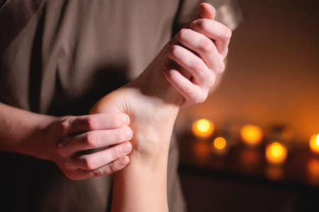 Close-up of a male masseur doing a leg and foot massage in a dark room against the background of candles professional massage in an office with dark lightの写真素材