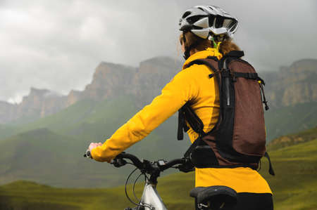 close-up young slender girl with a backpack and a bicycle helmet sits on a bicycle high in the mountains on a cloudy day and looks at the mountains and clouds.の写真素材