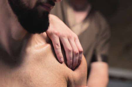close-up male masseur does a sports shoulder massage to a muscular male athlete in a room with a contrasting dark light. Professional sports massageの写真素材