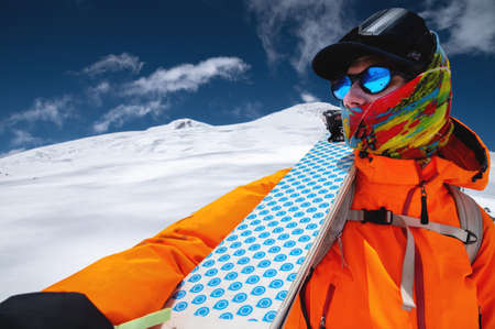 Portrait of a stern climber skier in sunglasses and a cap with a ski mask on his face. holds his skis on his shoulder and looks away against the backdrop of Mount Elbrusの写真素材