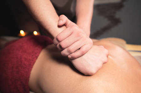 Young man masseur doing sports massage to a muscular athlete. Professional back massage in a dark room with candles. Relaxation and spa recovery of the muscular apparatusの写真素材