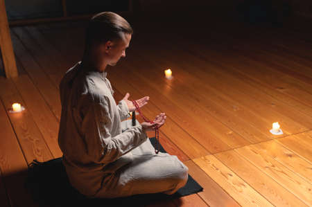 A man in clothes for practice and meditation sits in a lotus pose and holds red rosary to...