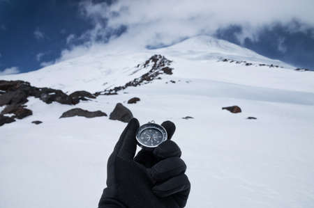 A metal magnetic compass in the hand of a traveler in a black glove against the backdrop of...
