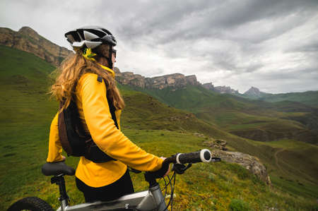 A slender girl cyclist with red hair in a helmet and with a backpack stands on a mountain bike high...