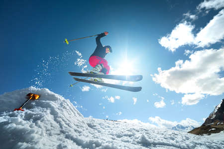 A young woman in sunglasses in tension jumps on skis from a springboard of snow against the blue sky and clouds. Uncertain performance of a trick on skisの写真素材