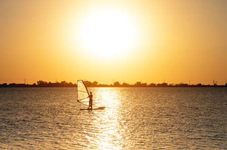 A beginner windsurfer woman stands on a board with a sail on a sunset background. Windsurfing schoolの写真素材