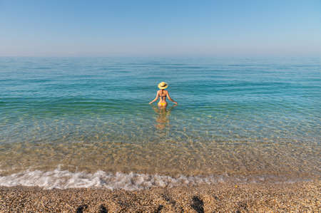 Attractive slender young Caucasian woman in a swimsuit and a straw hat stands waist-deep in sea water in the morning when there is haze over the seaの写真素材
