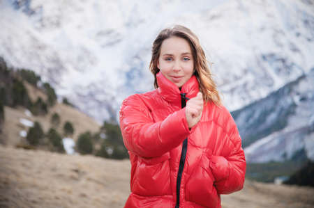 Portrait of a blonde young woman with bright blue eyes in a black dress and a red down jacket walks in the fall in the mountains. Walk in the highlandsの写真素材