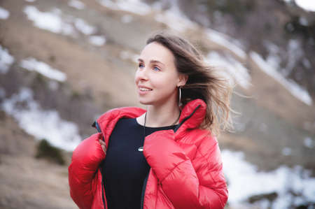Portrait of a blonde young woman with bright blue eyes in a black dress and a red down jacket walks...