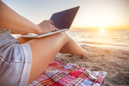 Close-up of a slender young woman on the beach at the sunset time by the sea works on her laptop holding him on her lap. Freelance concept for remote work and work while on vacationの写真素材