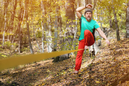 A bearded man in age balances while sitting on a taut slackline in the autumn forest. Outdoor...