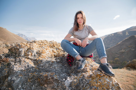 Attractive brooding young caucasian woman in gray T-shirt and jeans sits on a cliff against the mountains on a sunny dayの写真素材