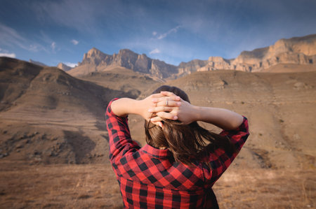 Young happy woman in a T-shirt and jeans stands high in the mountains with her arms outstretched enjoying the view of the mountains and the epic nature of the landscapeの写真素材