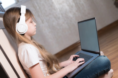 A smart little girl of seven years old in white headphones with a laptop in her hands is pushing on the floor in her room. The young generation on the Internet and IT technologyの写真素材