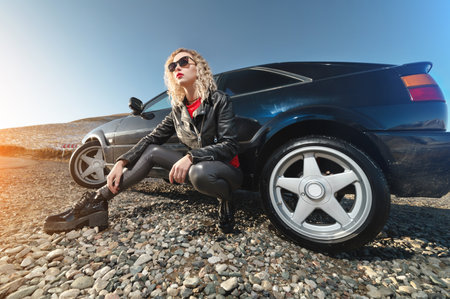 An attractive blonde in sunglasses and leather clothes sits near a black sports car on a country road in the mountains at sunset. Retro style 90sの写真素材