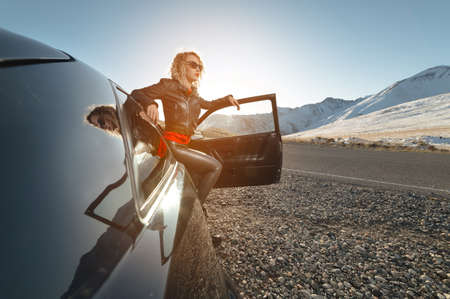 Fashion girl standing next to a retro sports car in the sun. Stylish woman in a leather suit is...
