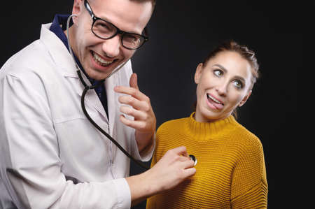 A young doctor with glasses listens to the heartbeat of a woman making faces. Medical humor. False diseasesの写真素材