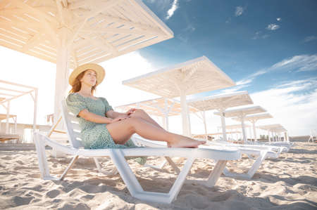 Young attractive caucasian woman in a green dress and a straw sits resting on a lounger under a wooden umbrella on a sandy beach of the seashore or ocean.の写真素材