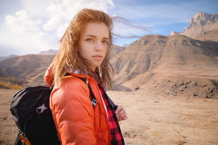 Young attractive Jewish woman in a down jacket and with a backpack against the backdrop of the epic mountains. Looks pensively at the cameraの写真素材