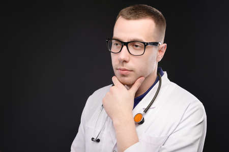 Portrait of unhappy tired young doctor with stethoscope with headache on dark background. Problems with the medicの写真素材