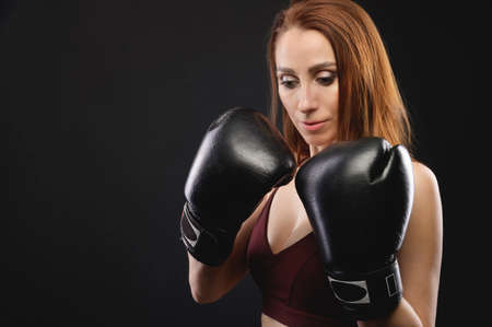Caucasian fitness girl in boxing gloves stands in a rack on a black background, portrait of a strong and independent woman fighterの写真素材