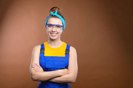 Attractive Caucasian young woman in work overalls and safety glasses against a brown background. Repair that will be done for you. Strong independent womanの写真素材